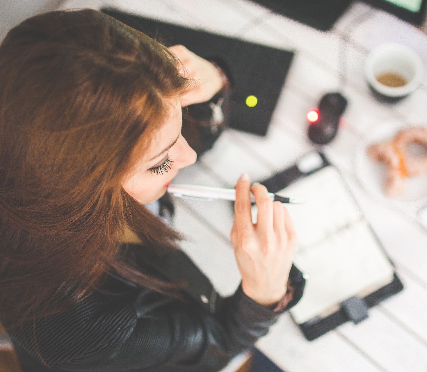 A woman with long red hair chewing her pen as she makes notes in a notebook